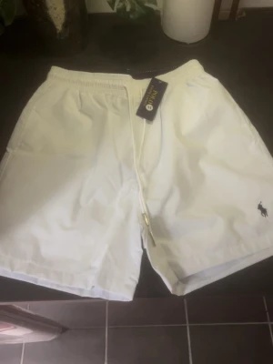 Vita shorts från Polo Ralph Lauren - Snygga vita shorts från Polo Ralph Lauren med elastisk midja och snörning. Klassisk logga broderad på benet och praktiska fickor både fram och bak. Perfekta för sommardagar och enkel att matcha med olika stilar.