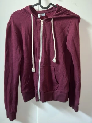 Vinröd hoodie med dragkedja H&M Divided - Vinröd hoodie från H&M Divided med vit dragkedja och vita snören i huvan. Modellen har två fickor framtill och ribbade muddar vid ärmslut och nederkant.