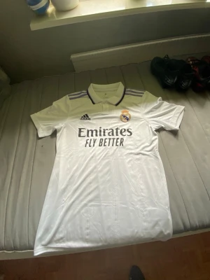 Real Madrid 22/23 matchtröja - Säljer en vit Real Madrid matchtröja från Adidas med klubbmärke broderat på bröstet och svart Adidas-logga. Tröjan har krage med knapp, lila detaljer på axlarna och trycket 'Emirates Fly Better' på framsidan. Perfekt för fotbollsfans!