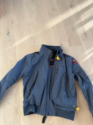 Grå vindjacka från Parajumpers - Snygg grå vindjacka från Parajumpers med gul detalj vid dragkedjan och patch på ärmen. Jackan har flera dragkedjeförsedda fickor, hög krage och meshfoder. Perfekt för dig som vill ha en stilren och funktionell jacka med coola detaljer.