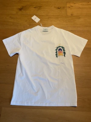 Vit Casablanca t-shirt med tryck - Säljer en vit t-shirt från Casablanca med färgglatt tryck av en byggnad och gröna blad på bröstet och ryggen. T-shirten har rund hals och korta ärmar. Perfekt för dig som gillar stilrena plagg med unika detaljer. Den har en liten svart fläck men den går nog lätt att ta bort