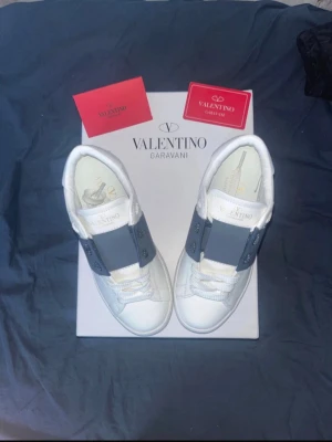 Valentino Garavani vita sneakers med mörk blå rem - Snygga vita sneakers från Valentino Garavani med bred mörk blå rem över vristen och coola nitar på sulan. Skorna är i läder och har en chunky sula som ger extra attityd. Perfekta för dig som vill ha en stilren men ändå unik look.