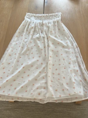 Vit långkjol med små blommor - Söt vit långkjol med små rosa blommor och smock i midjan med knytband. Kjolen har ett luftigt och lätt tyg som passar perfekt till varma dagar. Inga fläckar eller skador.