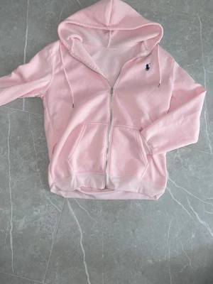 Ljusrosa hoodie från Ralph Lauren - Säljer en ljusrosa hoodie från Ralph Lauren med dragkedja framtill och två fickor. Klassisk huva och den ikoniska mörkblå loggan broderad på bröstet. Mjuk insida och ribbade muddar vid ärmar och nederkant. Perfekt för en avslappnad och stilren look.