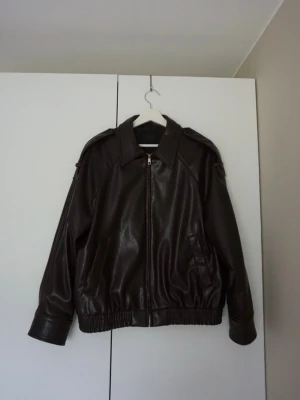 Mörkbrun skinnjacka från Zara - Snygg mörkbrun skinnjacka från Zara med dragkedja framtill, bred krage och resår i midjan. Jackan har axelklaffar och fickor på sidorna. Orginalpris 811 kr