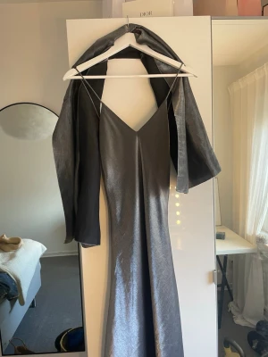 Silverfärgad långklänning med sjal - Elegant silverfärgad långklänning med smala axelband och v-ringning. Klänningen har en glansig finish och medföljer en matchande sjal i samma material. Perfekt för dig som vill sticka ut på nästa bal eller fest.