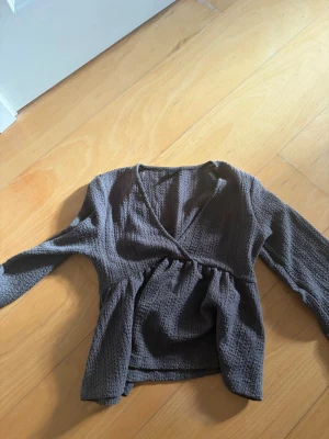 Mörkbrun omlottblus Gina Tricot - Säljer en mörkbrun omlottblus från Gina Tricot i storlek S. Blusen har lång ärm, v-ringad omlottfront och är gjord i ett mjukt, strukturerat tyg som ger en avslappnad men ändå stilren look. Perfekt att matcha med jeans eller kjol.