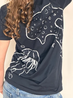 Svart t-shirt med vit grafisk print - Cool svart t-shirt med vit grafisk print av manet och havsinspirerat motiv. T-shirten har korta ärmar och en avslappnad passform. Allt är handmålat