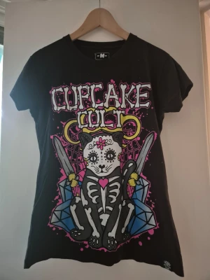 Svart Cupcake Cult t-shirt med kattmotiv stlM - Cool svart t-shirt från Cupcake Cult med stort färgglatt tryck av en skelettkatt, diamanter och svärd på framsidan. Texten 'Cupcake Cult' syns både fram och bak. T-shirten har rund hals och korta ärmar. Stl.M. Bra skick!