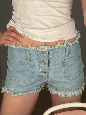 Ljusblå denimshorts med fransar - Säljer ett par ljusblå denimshorts med fransade kanter och stora framfickor. Shortsen har tre synliga knappar framtill. Perfekta för sommaren💕