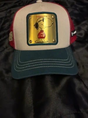 Peanuts x Goorin Bros keps Snoopy - Säljer en unik Peanuts x Goorin Bros keps med Snoopy-motiv på en guldfärgad patch framtill. Kepsen har vit front, röda mesh-sidor och mörkgrön böjd skärm med vita sömmar. Justerbar snapback och broderade detaljer på sidorna. Perfekt för dig som gillar streetwear och popkultur.