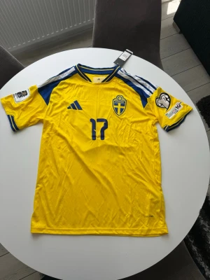 Sverige matchtröja Adidas #17 Gyökeres - Gul matchtröja från Adidas för svenska landslaget med blå detaljer, nummer 17 och namnet Gyökeres på ryggen. Tröjan har officiella landslagsmärket på bröstet och tryck på ärmarna. Perfekt för dig som vill stötta Sverige på matchdag!