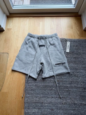 Grå Essentials Fear of God shorts - Snygga grå shorts från Essentials Fear of God med långa dragsnören och elastisk midja. Tillverkade i mjukt sweatshirtmaterial och har logga tryckt på vänster ben. Perfekta för en avslappnad och trendig stil.