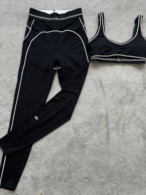 Svart träningsset från Alo - Snyggt svart träningsset från Alo med vita kontrastsömmar. Setet består av ett par tights med hög midja och en matchande sport-bh med breda axelband. Båda plaggen har Alo-logga och är gjorda i stretchigt funktionsmaterial som passar perfekt till träning.