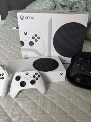 Xbox Series S med 3 handkontroller - Säljer en vit Xbox Series S med 512GB SSD och extra 1TB lagringsutrymme. Konsolen är kompakt och har en rund svart ventilationsyta. 3 trådlösa handkontroller ingår, 2 vit och en svart Elite-kontroll. Perfekt för gaming och smidig att ta med sig.