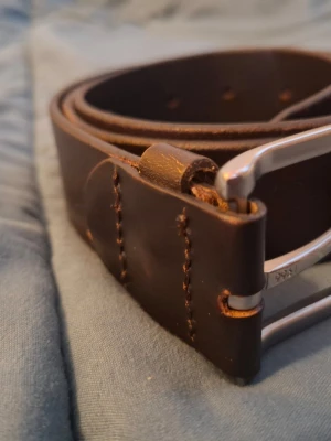 Tommy Hilfiger Bälte - Säljer ett stilrent och klassiskt bälte från Tommy Hilfiger i storlek 85 cm. Bältet är tillverkat i kraftigt, mörkt kvalitetsläder med en snygg inpräglad logotyp på sidan. Det har även den ikoniska Hilfiger-flaggan som en diskret detalj vid spännet.Ett perfekt herrbälte som passar lika bra till jeans som till kostymbyxor.