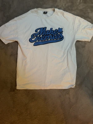 Vit Make Enemies t-shirt med blå tryck - Säljer en vit t-shirt från Make Enemies med ett stort blått tryck på bröstet. T-shirten har klassisk passform och korta ärmar, perfekt för dig som gillar streetwear och grafiska prints.
