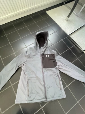 Grå vindjacka från Under Armour - Snygg grå vindjacka från Under Armour med huva och dragkedja. Jackan har en kontrasterande mörkgrå bröstficka med logga, två sidofickor med dragkedja och sportig design. Perfekt för blåsiga dagar och outdoor-aktiviteter.