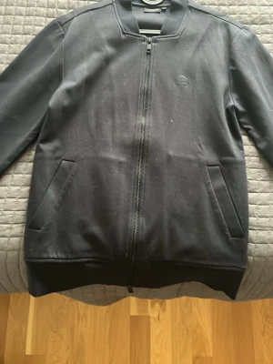 Svart ziptröja från J.Lindeberg - Snygg svart ziptröja från J.Lindeberg med diskret broderad logga på bröstet. Tröjan har två fickor framtill, ribbade muddar och en stilren ståkrage. Tillverkad i mjukt bomullsmaterial som känns bekvämt hela dagen. Tröjan är använd fåtal gånger och pris kam diskuteras och tar emot byten!