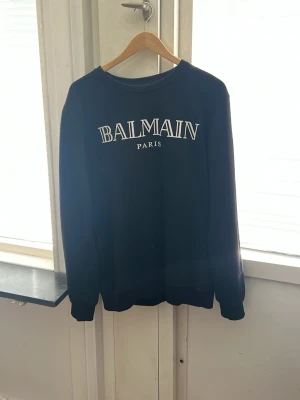 Svart Balmain hoodie med tryck - Snygg svart hoodie från Balmain med stort vitt Balmain Paris-tryck på bröstet. Klassisk modell med rund halsringning och långa ärmar. Perfekt för dig som gillar streetwear och exklusiva märken. Passar även dem som har storlek S