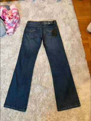 Blå bootcut-jeans med detaljer - Snygga blå bootcut-jeans från Sexy Girl med dekorativa detaljer i form av strass på bakfickorna. Klassisk femficksmodell med hällor för skärp och låg midja. Perfekta för dig som gillar en tidlös och trendig look.