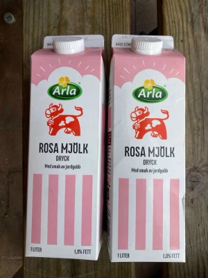 Tom Arla Rosa Mjölk jordgubb 1L - Två tomma förpackningar Arla Rosa Mjölk dryck med smak av jordgubb. Kartongerna är vita och rosa med en röd ko på framsidan och grönt Arla-märke. Varje förpackning innehåller 1 liter och har 1,0% fett.