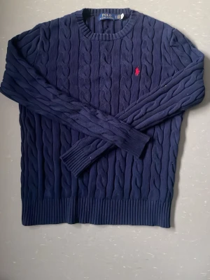 Mörkblå kabelstickad tröja Ralph Lauren - Kabelstickad mörkblå tröja från Ralph Lauren med rund halsringning och röd broderad logga på bröstet. Tröjan har långa ärmar och ribbade muddar vid ärmslut och nederkant. Perfekt för lager på lager och klassisk stil. Original pris: 2595kr 