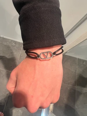 Valentino armband  - Snyggt svart armband från Valentino med dubbla band i textil och en silverfärgad metallbricka med det ikoniska V-logotypen. Enkel och stilren design som passar perfekt till din vardagsstil.
