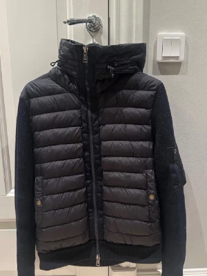 Svart Moncler cardigan  - Snygg svart dunväst från Moncler med stickade ärmar och vadderad framsida. Västen har en stor huva med dragsko, två fickor med tryckknappar och dragkedja framtill. Moncler-logga på ärmen och cool mix av material för en modern look. 