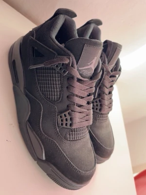 Svarta Air Jordan 4 sneakers/Black cats - Snygga svarta Air Jordan 4 sneakers med ikonisk Jumpman-logga på plösen. Skorna har meshdetaljer på sidorna, tjock sula och klassiska plastdetaljer vid snörningen. Ovandelen är i mockaliknande material och designen är chunky och sportig.