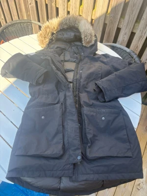 Svart parkasjacka från Everest - Snygg svart parkasjacka från Everest med stor huva och avtagbar fuskpäls. Jackan har två stora framfickor med lock och tryckknappar, dragkedja och vindslå framtill. Perfekt för kalla dagar med sitt vadderade material och klassiska design.
