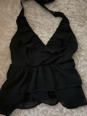 Svart volang topp med knyt i nacken - Snygg svart topp med volangdetaljer och djup v-ringning. Toppen har knyt i nacken och öppen rygg. Använd ett fåtal ggr💗