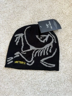 Svart mössa från Arc'teryx - Svart mössa från Arc'teryx med grått grafiskt skelettmönster och gul broderad logga framtill. Tunn och smidig modell i mjukt material, perfekt för kyliga dagar. Snygg och stilren design som passar till det mesta.