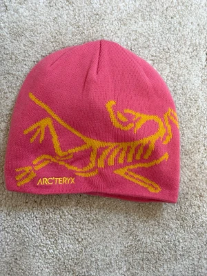 Rosa mössa från Arc'teryx - Snygg rosa mössa från Arc'teryx med ett gult dinosaurie-skelettmotiv och broderad logga framtill. Mössan är stickad och har en klassisk, rundad form. Perfekt för kalla dagar och outdoor-aktiviteter.