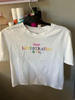 Vit t-shirt från Levi's med brodyr - Vit t-shirt från Levi's med färgglad broderad logga på bröstet. Klassisk rund hals och korta ärmar. Tillverkad i mjuk bomull och har en avslappnad passform. Perfekt för en trendig och enkel stil.
