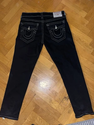 Svarta True Religion jeans med kontrastsömmar - Snygga svarta jeans från True Religion med vita kontrastsömmar och ikoniska fickdetaljer bak. Klassisk femficksmodell med raka ben och knappgylf. Perfekta för dig som gillar streetwear och vill sticka ut med detaljer.