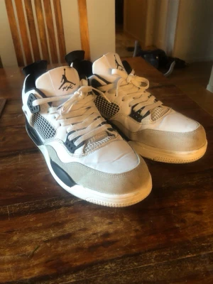 Air Jordan 4 Retro Vit/Svart/Grå - Snygga Air Jordan 4 Retro sneakers i vitt, svart och grått. Skorna har klassisk mesh på sidorna, Jumpman-logga på plösen och hälen samt detaljer i mocka och läder. Perfekta för dig som gillar streetwear och vill sticka ut med en ikonisk siluett.