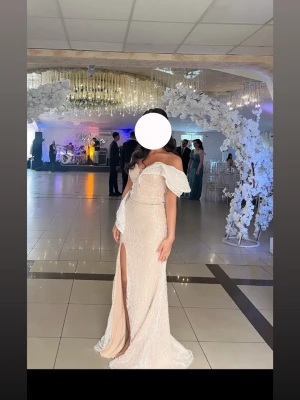 Lång offshoulder klänning i champagne - Elegant långklänning i champagnefärg med offshoulder-ärmar och slits framtill. Klänningen har volangdetaljer och en figurnära passform, perfekt för speciella tillfällen. Materialet har en lätt glittrig yta som ger en lyxig känsla.