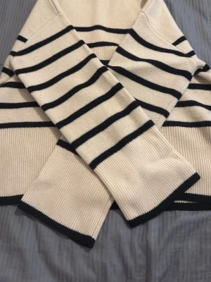 Randig beige stickad tröja - Snygg stickad tröja i beige med svarta horisontella ränder. Tröjan har långa ärmar och ribbade muddar samt en rak passform. Perfekt för dig som gillar klassisk och tidlös stil. Materialet känns mjukt och bekvämt.