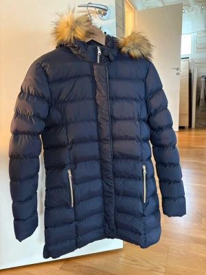 Mörkblå lång dunjacka med fuskpäls - Snygg mörkblå dunjacka med huva och fuskpälsdetalj. Jackan är lång, har två dragkedjefickor framtill och quiltad design. Perfekt för kalla vinterdagar och riktigt varm. Stängs med dragkedja och tryckknappar.