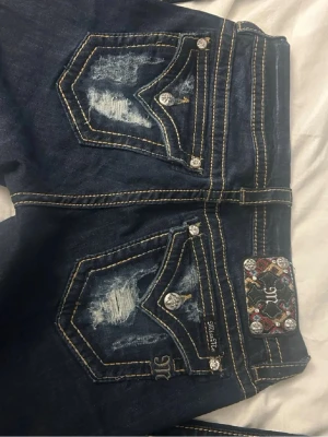 Mörkblå Miss Me jeans med detaljer bootcut lowwaist - Snygga mörkblå jeans från Miss Me med slitna detaljer på bakfickorna, kontrastsömmar och dekorativa knappar. Bakfickorna har broderade mönster och glittriga stenar. Klassisk femficksmodell med raka ben och låg midja. Storlek 28. Innerbenslängd 90 cm. Midjemått rakt över 37 cm.