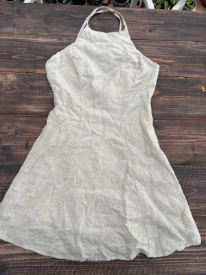 Beige halterneck klänning i linne - Snygg beige halterneck klänning i linne med figurnära passform och utsvängd kjol. Perfekt för varma dagar och enkel att styla med accessoarer. Klänningen har en minimalistisk design utan synliga detaljer eller mönster. Dubbeltyg.