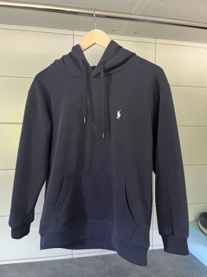 Mörkblå hoodie från Polo Ralph Lauren - Mörkblå hoodie från Polo Ralph Lauren med klassisk huva och dragsko. Har en vit broderad logga på bröstet och en stor känguruficka framtill. Tillverkad i mjuk bomullsblandning och har ribbade muddar vid ärmslut och nederkant. Den är väldigt liten i storleken så hör av dig vid funderingar. Det står att den är i storlek S men de skulle passa bättre för dig som har storlek XS till XXS.