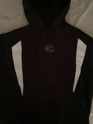 Svart Nike Air hoodie med vita detaljer - Snygg svart hoodie från Nike Air med vita paneler längs axlarna och bröstet. Klassisk huva och Nike Air-logga framtill. Perfekt för en sportig och avslappnad stil. Materialet känns mjukt och bekvämt mot huden