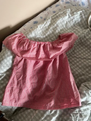 Rödvit offshoulder randig topp - Snygg offshoulder topp med röda och vita ränder. Toppen har en volang runt axlarna och är ärmlös, perfekt för varma dagar. Tillverkad i ett lätt bomullstyg som känns skönt mot huden. Passar dig som gillar en avslappnad men trendig stil.