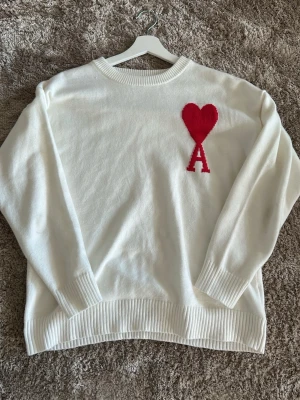 Ami Paris - Vit stickad tröja från Ami Paris i helt nytt skick med rund halsringning och ribbade muddar. På bröstet finns det ikoniska röda hjärtat med bokstaven A. Tröjan har en avslappnad passform och är tillverkad i mjukt material.