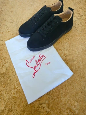 Svarta sneakers från Christian Louboutin - Snygga svarta sneakers från Christian Louboutin med klassisk röd sula och diskret logga. Skorna har lågt skaft, svarta skosnören och ovandel i mocka. Perfekta för dig som vill ha en clean och exklusiv look. Oanvända storlek 44.