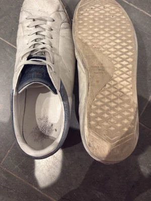 Vita Louis Vuitton sneakers - Säljer ett par vita sneakers från Louis Vuitton med vit snörning och blå detalj vid hälen. Skorna har klassisk låg profil och ovandel i läder. Snygg och stilren design som passar till det mesta. Yttersulan har diskret mönster för bra grepp. Nypris ungefär 10000 men slitna därför billigt pris
