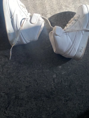 Vita high top sneakers från FILA - Säljer ett par vita high top sneakers från FILA med klassisk design. Skorna har snörning, vadderad plös och krage samt ovandel i strukturerat läder. Perfekta för dig som gillar sportig och clean stil.