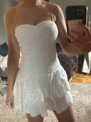 Vit kort klänning med spetsdetaljer - Supersöt vit kort klänning med smockad överdel och tunna axelband. Kjolen har volanger och broderade spetsdetaljer som ger en romantisk känsla. Perfekt för varma dagar och somriga tillfällen.